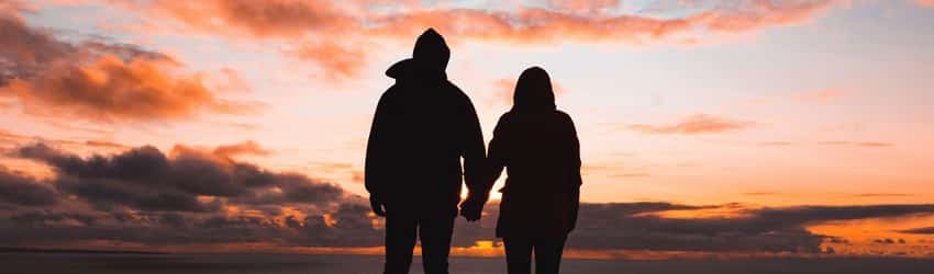 A silhouetted couple holds hands while watching the orange and purple sunset.