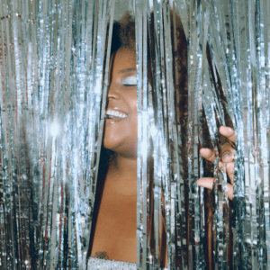 A black woman closes her eyes in pleasure as she enters through a shiny, glittery silver door covering.