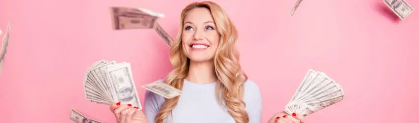 A smiling blonde woman holds up handfuls of money while cash flies around her.