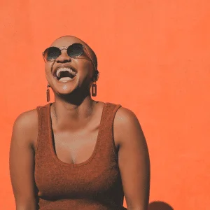 joyful woman with sunglasses and trendy earrings smiling against an orange backdrop