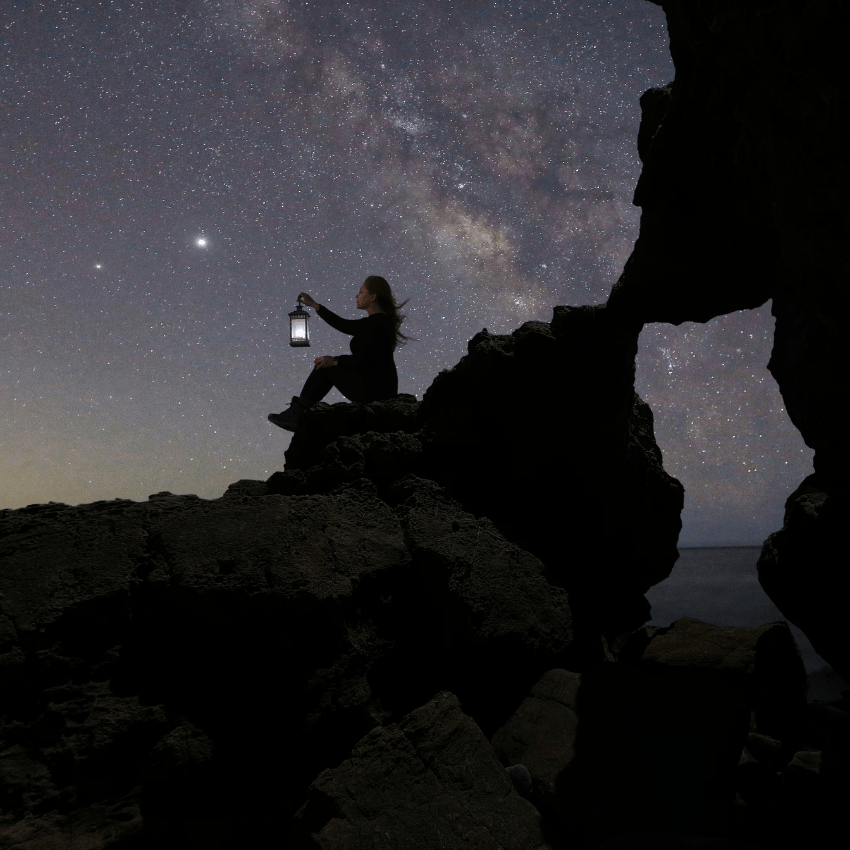 woman sitting on a rock with a lantern gazing at a purple starry night