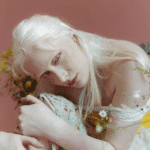 A girl with white hair and skin crouches over her knee holding a flower.