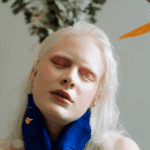 A woman with pale skin and white hair has her eyes closed. She wears a blue scarf around her neck.