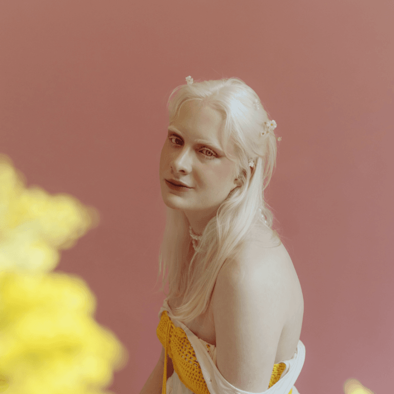 A pale woman with white hair sits looking at the camera. A yellow flower is blurred in the left corner.