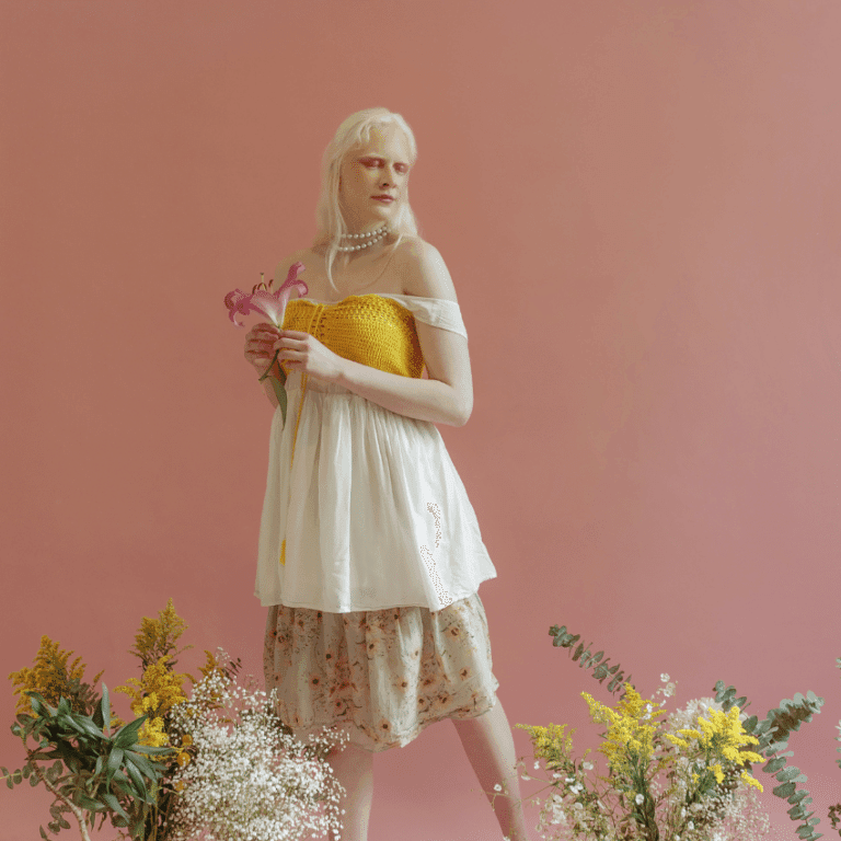 A woman with pale skin and white hair stands amongst fresh cut flowers